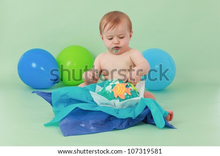 Brunette hair blue eyed baby boy looking down at his  ocean themed birthday party cupcake with orange fish and seaweed on it while sitting on green background with green and blue balloons behind him.