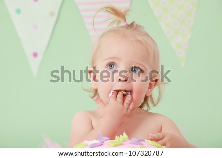 Blond hair blue eyed baby girl with pony tail eating and tasting vanilla sponge cake with pink and purple fondant icing while sitting on green background with pink green white and blue flag bunting