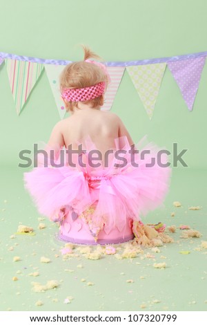 Adorable cheeky blond haired baby girl in tutu with pink head band in hair sitting on vanilla sponge cake with pink and purple hearts on icing with green seamless background and flag bunting behind