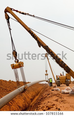 Large yellow side boom pipe layer or stringer industrial machine on orange dusty sandy outdoor construction site laying a pipe in a trench watching by workers