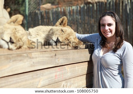 Young pretty brunette woman wearing grey and blue clothes standing by two beautiful sleeping wild lion cub stroking fur and smiling outside in nature park