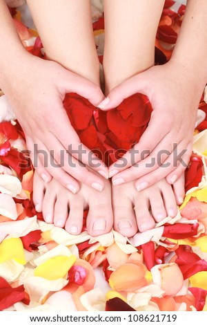 Clean and tidy well manicured soft female hands and feet resting on a bed of red yellow and orange petals while making a heart shape with her hands