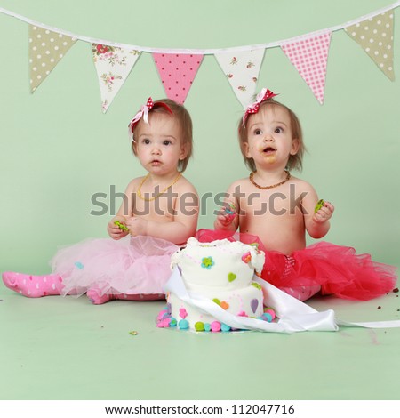 Two baby girl identical twin sisters sitting on green seamless background behind decorated birthday cake wearing red and pink tutus and bunting flags in the background about to break and smash cake