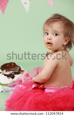 Baby girl sitting on green seamless background behind decorated birthday cake wearing red tutu and bunting flags in the background about to break and smash cake