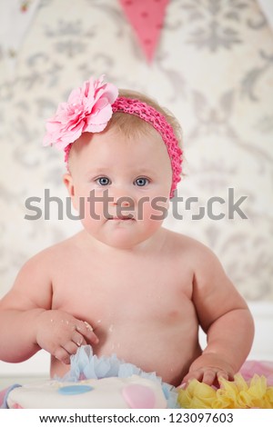 Cute happy blond baby girl in pink and yellow tutu and flower head band sitting on vintage wooden floor and background by smashed double tier polka dot decorated pink blue and white fondant iced cake