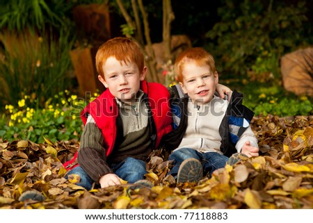 Two happy and smiling brothers or sons are sitting and hugging in a pile of colorful yellow and brown autumn / fall leaves in a garden or park setting