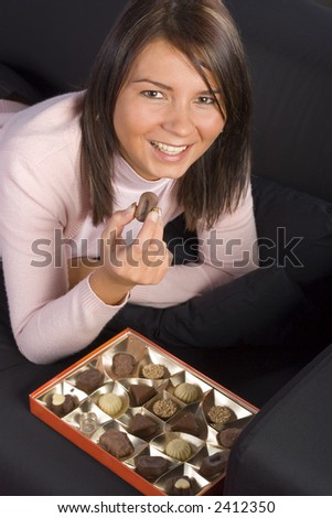Young woman lying on the black sofa with box of chocolates; Keeping chocolate in her hand.