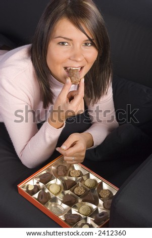 Young woman lying on the black sofa with box of chocolates; Putting chocolate to the mouth.