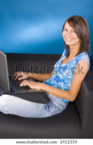 Young woman sitting on the black sofa with laptop. Blue background, in studio.