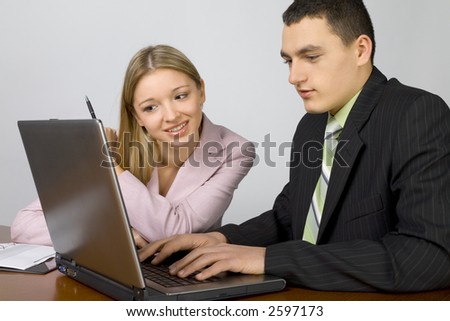 Man and woman at the office desk. There's laptop and notepad on it. Man's typing. Woman's pointing at the screen.