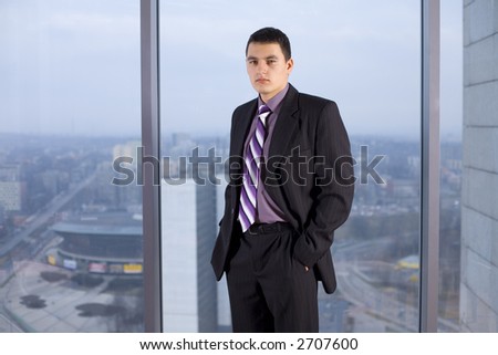 Businessman Standing and Looking at the Camera. There\'s Big Window With Big City View Behind Him.