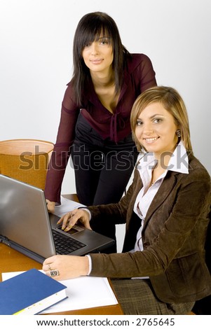 Women are next to the conference\'s table. Both are looking at the camera with smile. There\'s laptop on the table.