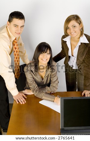 Two women and a man are at the desk. One of the women\'s sitting. They are all smiling to the camera.