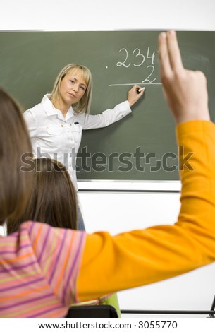 Mid age woman (teachear) are standing with chalk in hand close to greenboard. Two girls (class) are sitting opposite her. One of girls holding arm up. Focus on teacher\'s face.