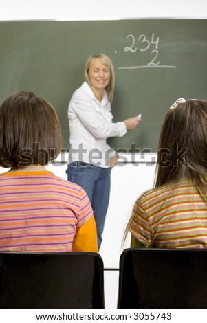 Woman (teachear) are standing with chalk in hand close to greenboard. Two girls (class) are sitting opposite her.  Focus on girl\'s backs. Teacher unfocus.