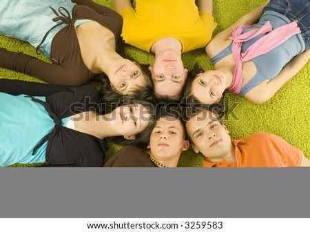 Group of 6 teenagers lying on the floor head next to head. They\'re looking at camera.