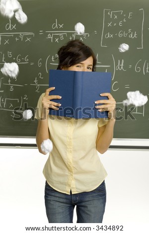 Teenage girl standing in classroom, in front of blackboard. Hiding behind  book. Someone throwing pieces of paper into her. Front view