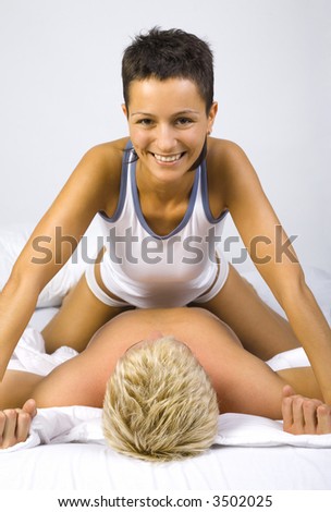 Young, smiling woman sitting on man in bed, showing her domination. Gray background, front view