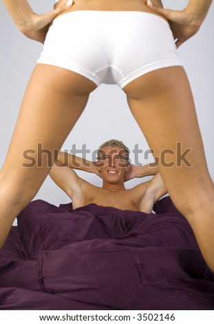Smiling young man, lying in bed in front of sexy woman. Gray background