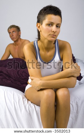 Young sad woman sitting on bed, nearby unsatisfied man. Woman looking at camera. Gray background, front view