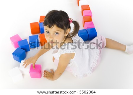 Little girl sitting on the floor and playing colored blocks. Looking at camera, white background. High angle view
