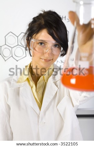 Young, beauty chemist girl in white apron. Holding beaker with orange liquid. Wearing goggles. Looking at camera. Gray background