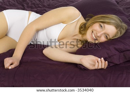 Young, sleepy, beautiful woman lying in bed on violet coverlet. Smiling, looking at camera