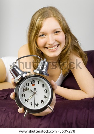 Young, beautiful woman lying in bed on violet coverlet.  Holding the clock and smiling. Focus on face and clock. Gray background. Looking at camera