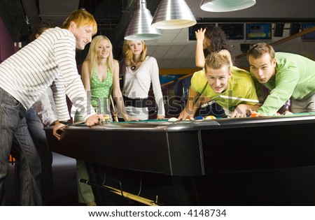 Large group of teenagers standing at pool table. Smiling nad looking at two teenage boys which ones playing billard.