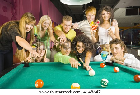 Large group of teenagers standing at pool table. Smiling and showing something. One person is playing billard