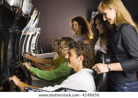 Large group of teenagers in amusement arcade. Teenage girls are standing and smiling, boys driving cars. Side view