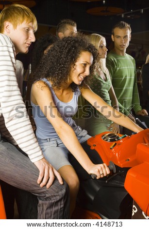 Large group of teenagers in amusement arcade. One teenage girl sitting on motorcycle. Rest of people are standing and smiling behind her