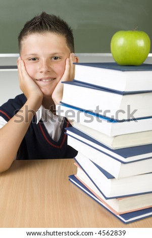 Young smiling boy sitting at desk nearby books. Apple lying on the books. Boy is looking at camera. Front view