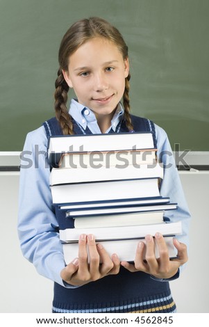 Young, smiling girl holding books. Standing in front of blackboard. Looking at camera, front view