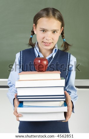 Young, smiling girl holding books. On books is lying apple. Standing in front of blackboard. Looking at camera, front view