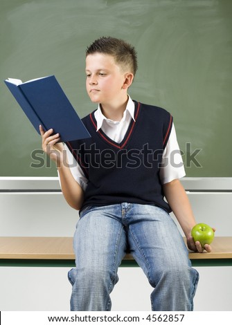 Young boy sitting on desk in front of blackboard. Holding book in one hand and reading. In other hand holding apple. front view