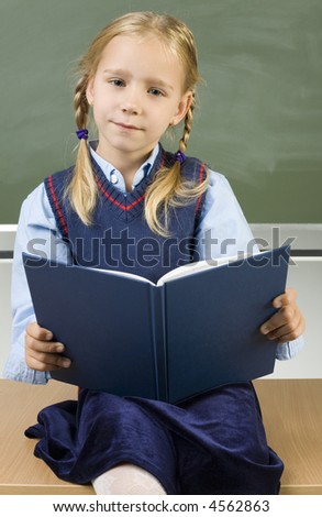 Little girl sitting on desk in front of blackboard. Holding book and smiling. Looking at camera. Front view