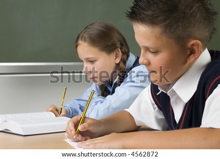 Young girl and boy sitting at desk and writing something. Boy is smiling. Side view