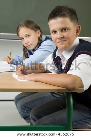 Young girl and boy sitting at desk and looking at camera. Holding pencils and smiling. Side view