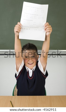 Young, smiling boy sitting at dest in front of blackboard with test in hand. Smiling and looking at camera. Hands up, front view