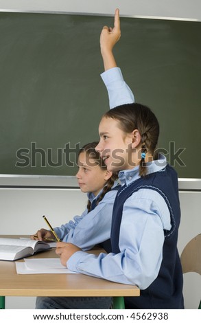 Girl holding her hand up. She wants to answer the teacher\'s question. There\'s another girl next to her. They\'re both sitting at the table. Blackboard behind them.
