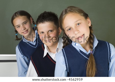 Portrait of three students: two girls and a boy in the middle of them. They're looking at camera and smiling. Blackboard behind them.