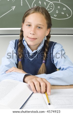 Young, smiling girl sitting at desk in front of blackboard over books. Holding pencil and looking at camera. front view