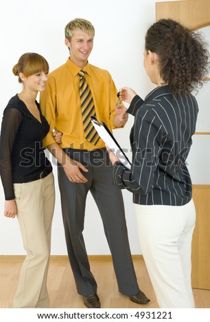 Three people standing in flat. One couple standing in front of camera and one woman standing back. Woman is giving keys to the man