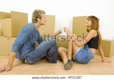 Young couple sitting on the floor in flat. They\'re looking happy. Celebrating removal with champagne and having toast. Side view, whole body