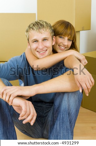 Young couple sitting on the floor in flat. They're looking happy. Woman is hugging man. They're looking at camera. Front view