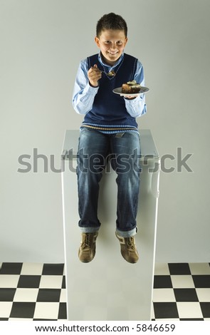 Happy boy sitting on fridge and eating cake. Front view. Grey background.