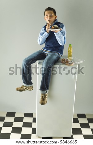 Young boy sitting on fridge and eating cake. He's delighting a taste of cake. Front view.