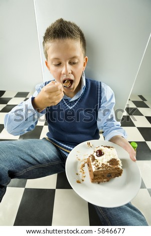 Young boy eating cake. He's sitting on the floor near by the fridge.