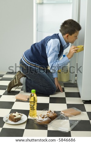 Young boy kneeling in front on the fridge and looking for something inside. Side view